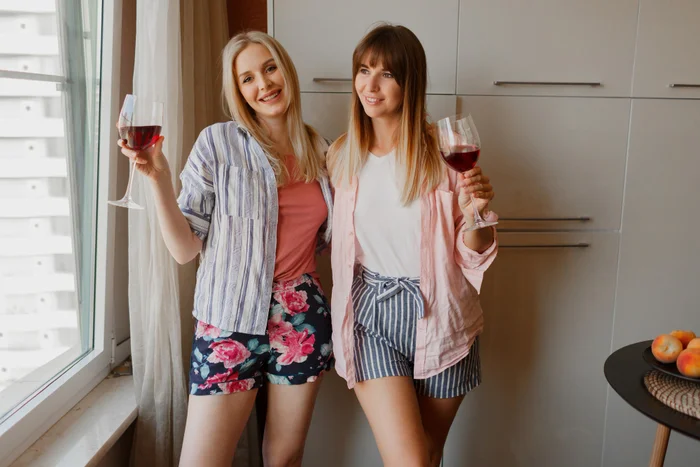 Couple of happy carefree women looking at window and holding glass of wine. cozy home atmosphere.