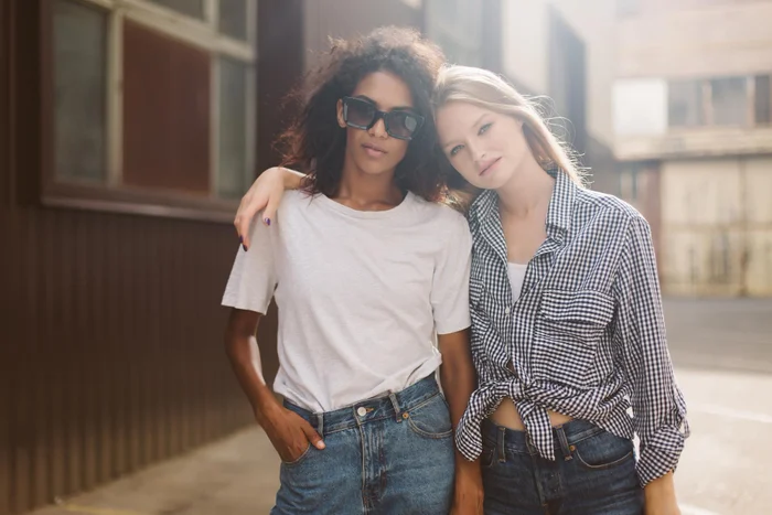 Young african american woman with dark curly hair in sunglasses and tshirt and pretty woman with blond hair in shirt dreamily looking in camera while spending time together